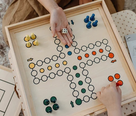 Zwei Personen spielen das Spiel 'Mensch ärgere Dich nicht'. Eine Person hält einen Würfel, die andere bewegt einen roten Spielstein. small foot Spielesammlung, 20 Klassiker.