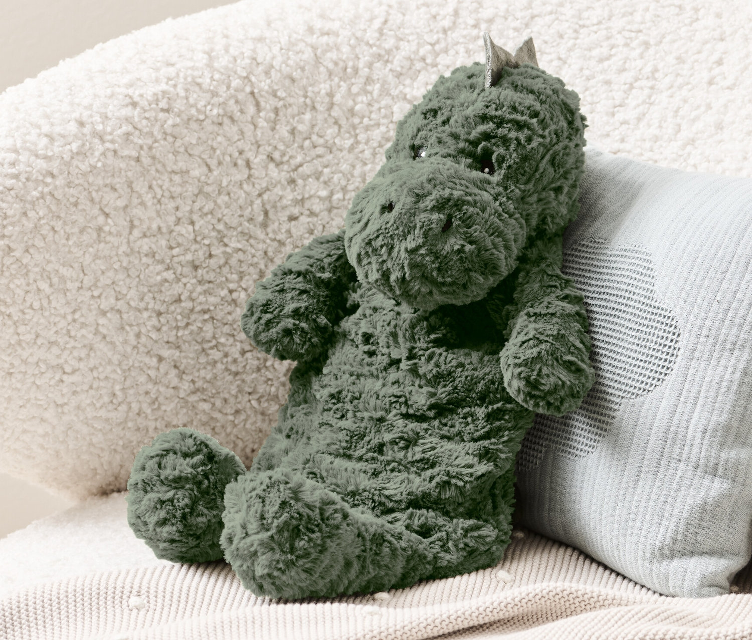 Eine grüne Dino-Wärmflasche sitzt auf einem cremefarbenen Sessel neben einem hellblauen Kissen.