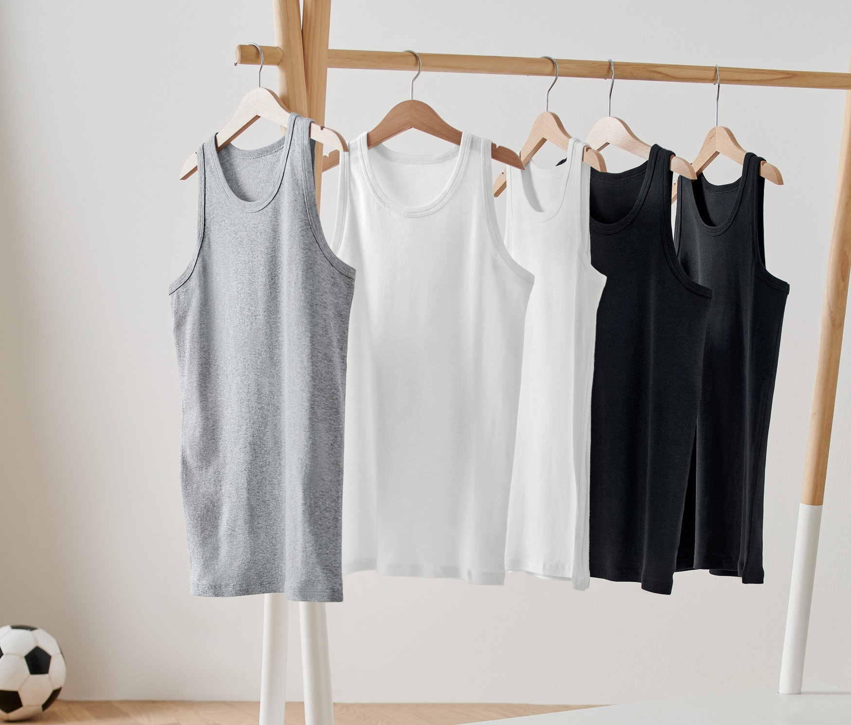Fünf Kinder-Unterhemden in Grau, Weiß und Schwarz hängen auf einem Holz-Kleiderständer. Im Hintergrund ist ein Fußball zu sehen.