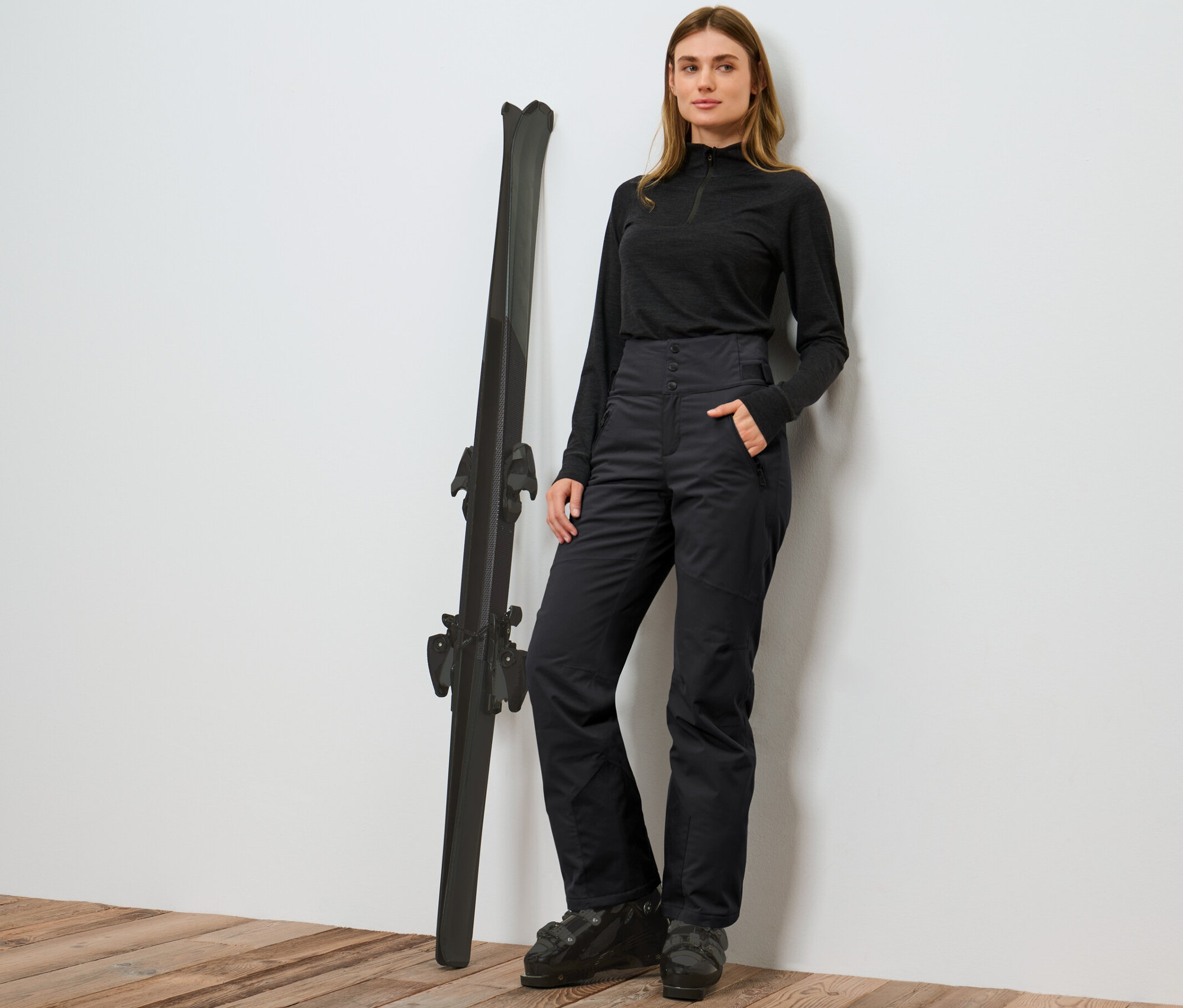 Frau posiert neben Skiern in einer schwarzen High-Waist-Skihose.