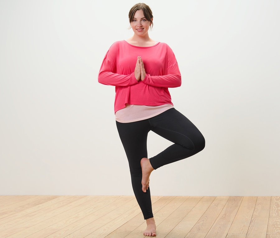 Frau im pinken 2-in-1-Sportshirt und schwarzer Sporttight steht in Baum-Pose.