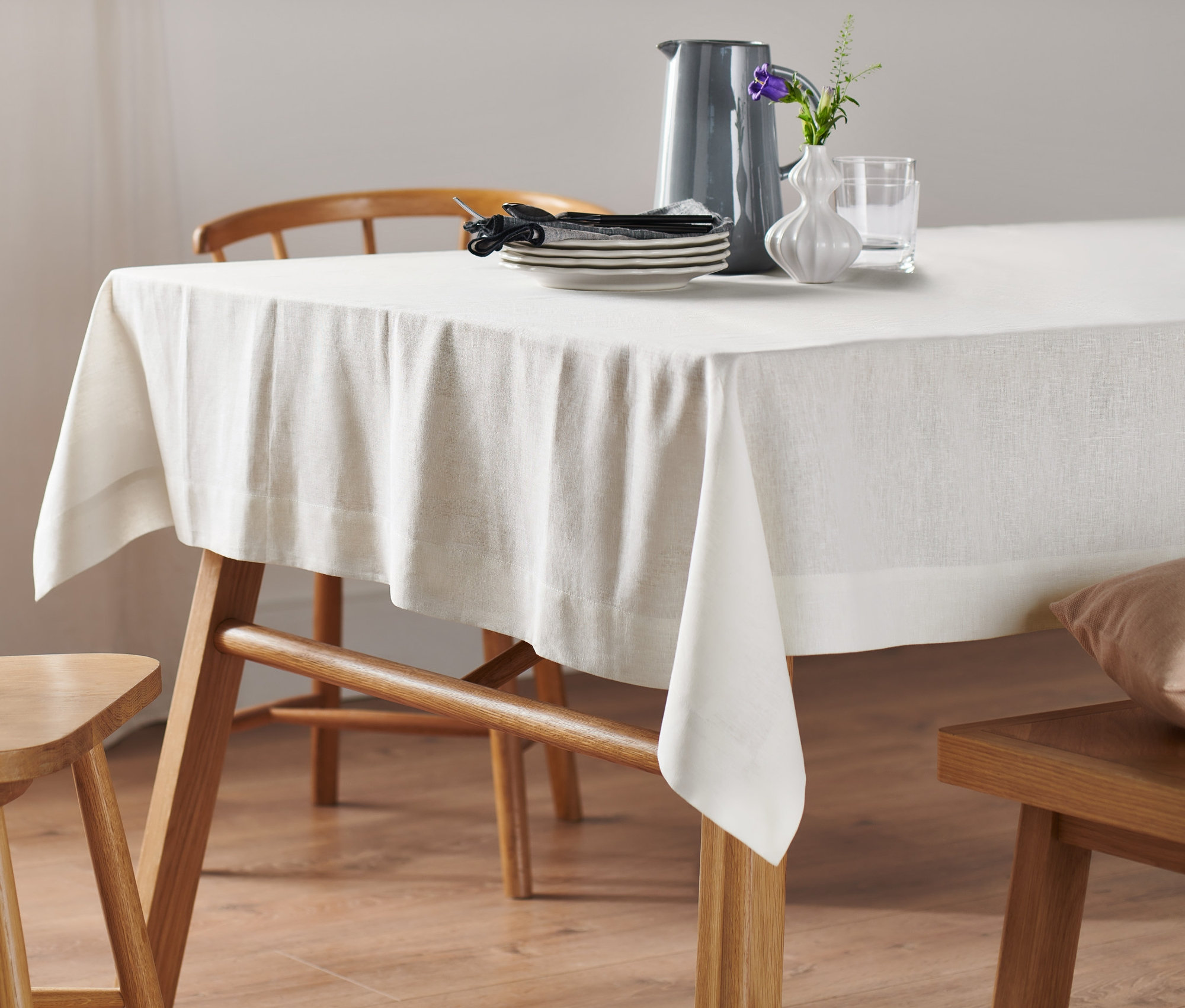 Eine normalgroße, weiße Leinen-Tischdecke liegt auf einem Holztisch. Auf der Tischdecke stehen eine Vase und Geschirr.