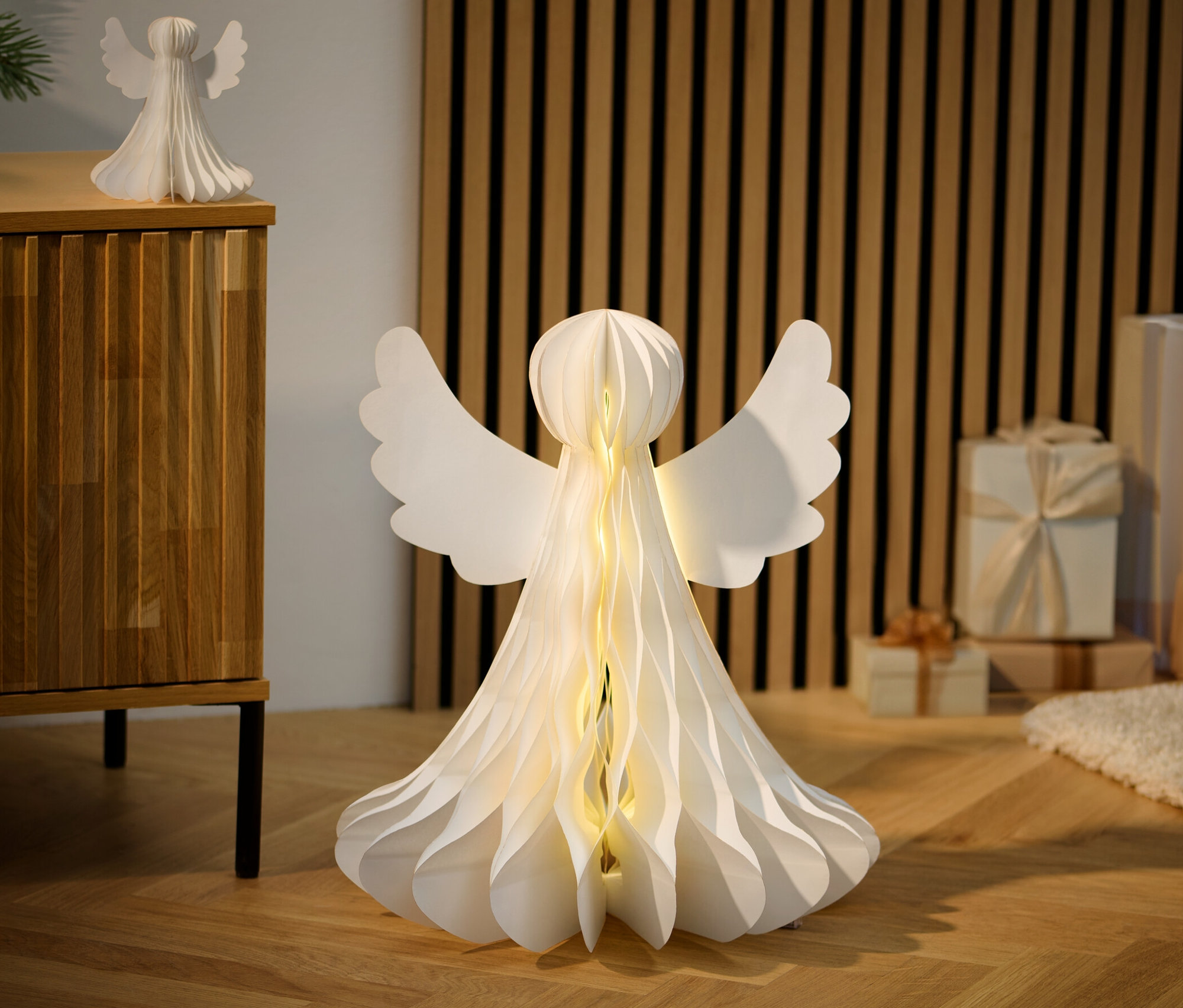 Zwei LED-Papier-Engel, XL, stehen auf einem Holzboden neben Geschenken.