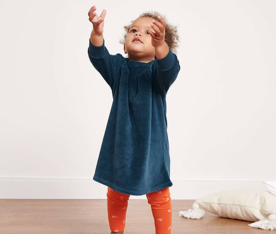 Ein kleines Mädchen greift nach Seifenblasen. Es trägt ein Baby-Nickikleid und orangefarbene Leggings.