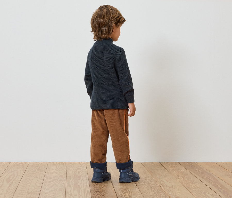 Ein Junge steht mit dem Rücken zur Kamera und trägt einen dunkelblauen Pullover, braune Hosen und blaue Stiefel.