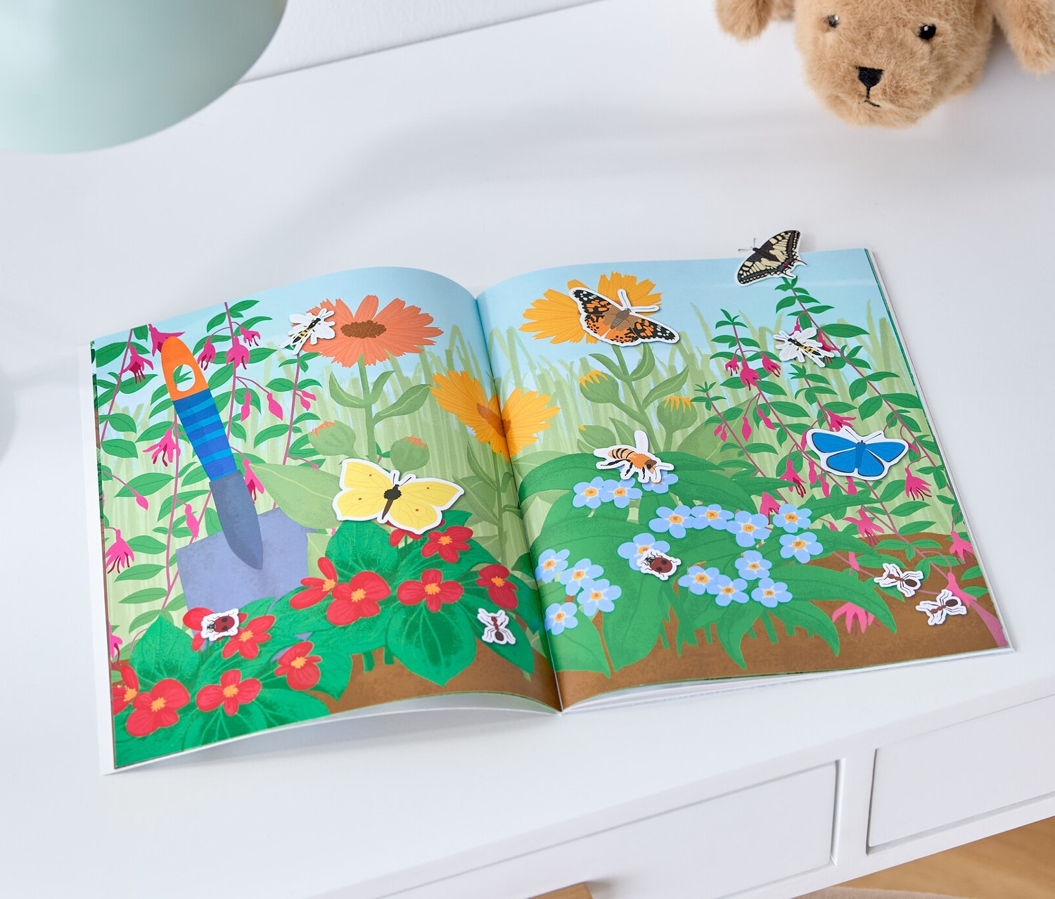 Auf einem weißen Tisch liegen ein geöffnetes Mein Stickerbuch »Jahreszeiten«, eine Lampe und ein Plüschhund.