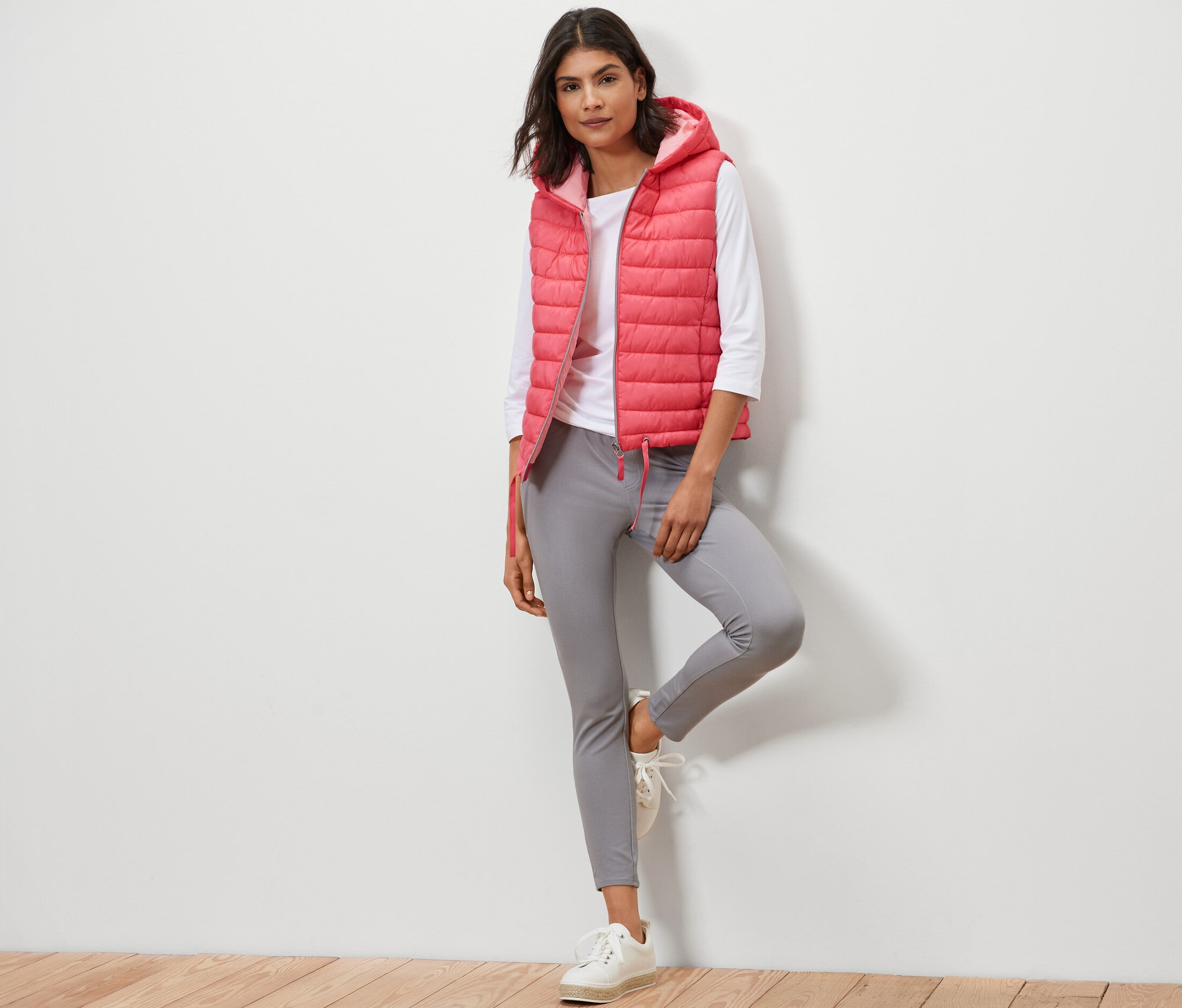 Frau posiert in Steppweste mit Kapuze, weißem Shirt und grauer Hose.