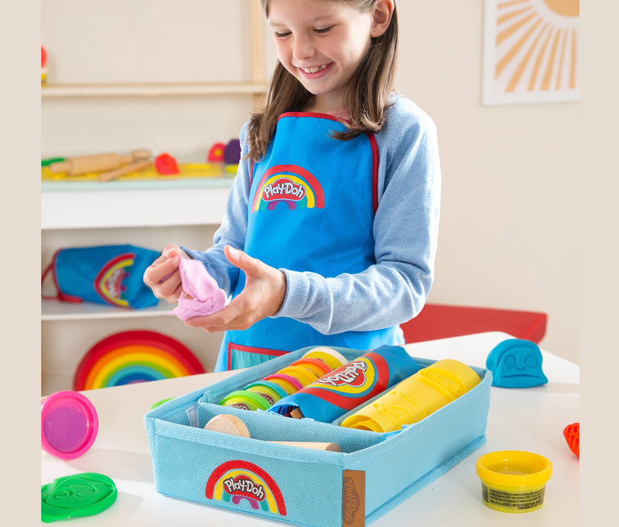 Ein Mädchen mit Schürze spielt mit Knete. Im Vordergrund befindet sich eine blaue Aufbewahrungsbox mit roba x Play-Doh Knet-Spielset 2.