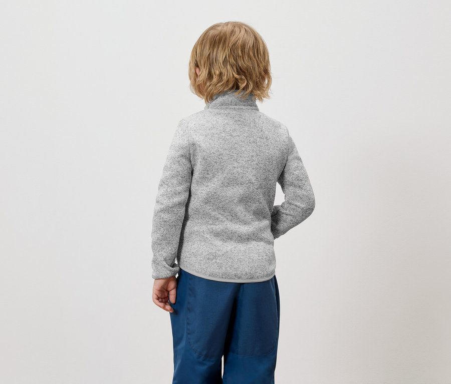 Ein Junge mit blonden Haaren steht mit dem Rücken zur Kamera und trägt einen grauen Pullover und blaue Hosen.