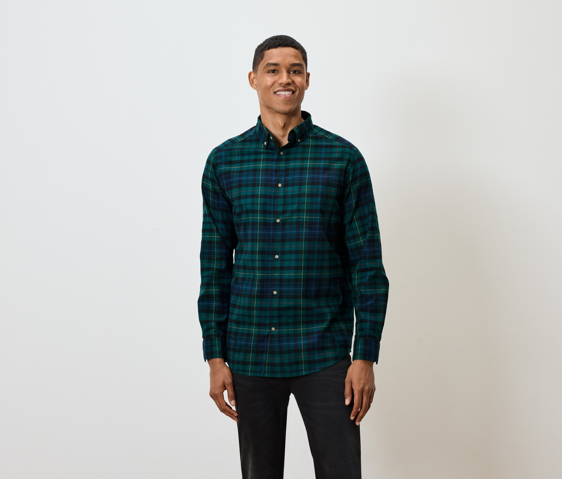 Ein kurzhaariger Mann posiert vor einem weißen Hintergrund und trägt ein grün-blau kariertes Langarmhemd und schwarze Jeans.