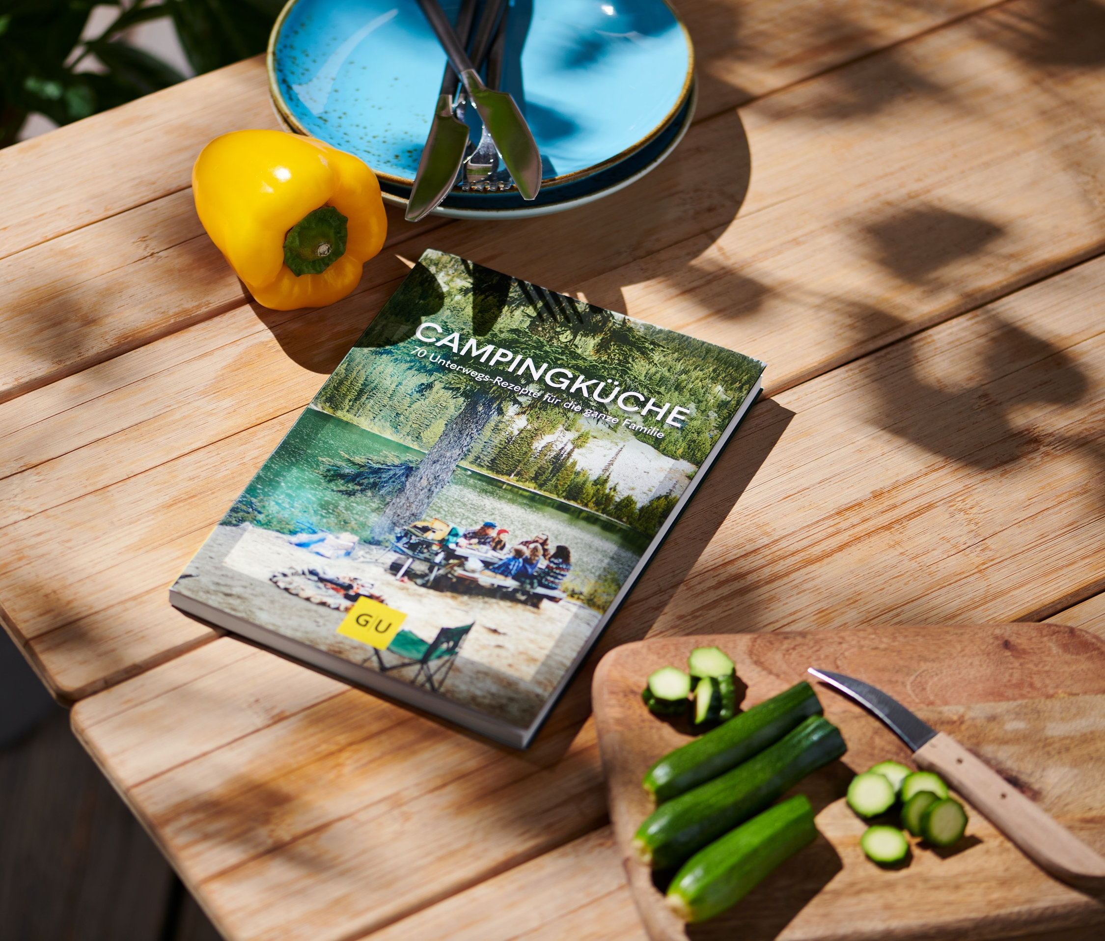 Auf einem Holztisch liegen das Buch "Campingküche", eine gelbe Paprika, blaue Teller mit Besteck und geschnittene Zucchini mit einem Messer.