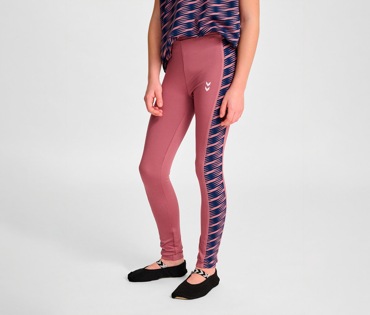 Mädchen posiert in rosa HUMMEL HMLJR Mix Mid Waist Tight Leggings mit blauen Streifen an der Seite und schwarzen Ballerinas.