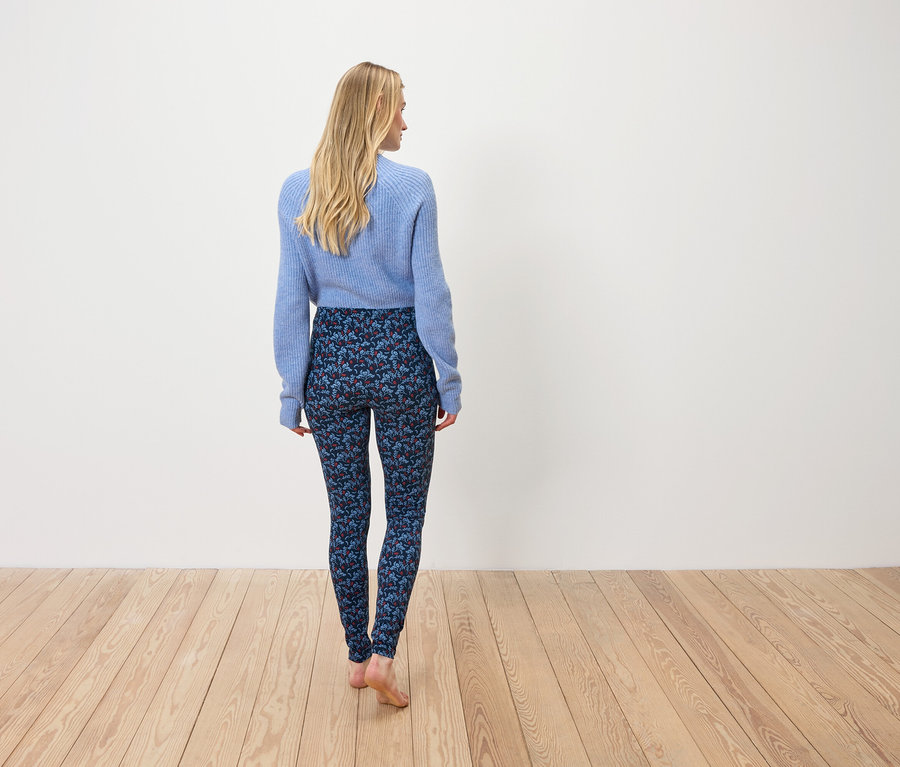 Frau in blauem Pullover und gemusterter Leggings steht mit dem Rücken zur Kamera.