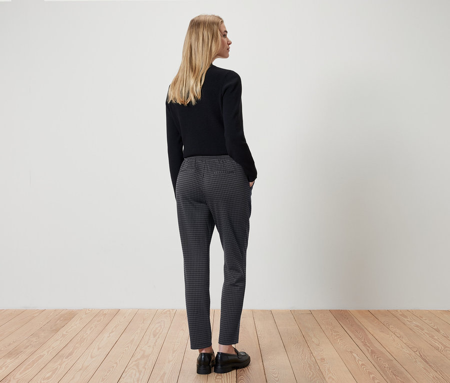 Frau steht mit dem Rücken zur Kamera und trägt ein schwarzes Oberteil und eine grau karierte Hose.