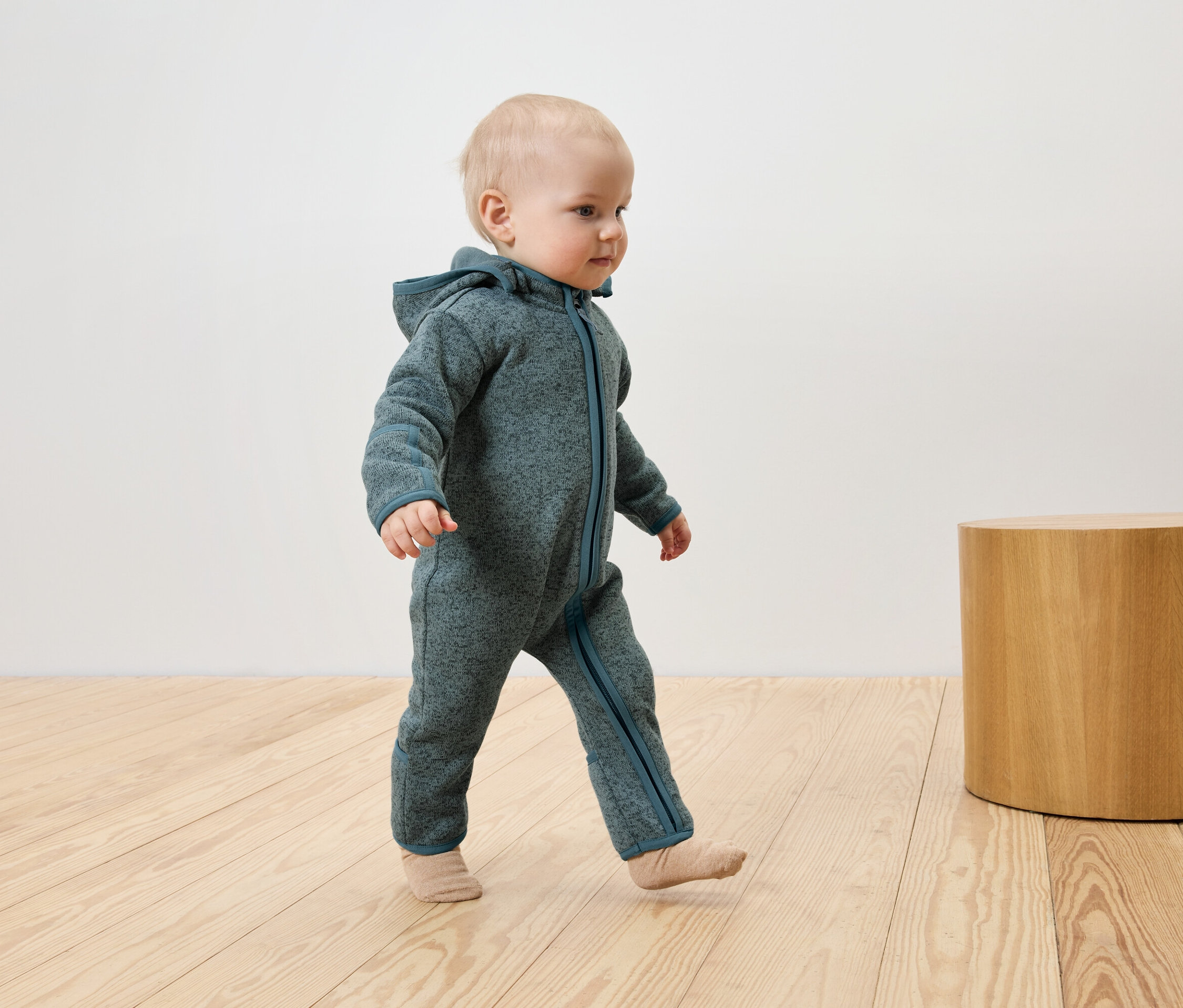 Kleinkind in blaugrünem Overall mit Kapuze geht auf Holzboden neben einem Holzhocker.