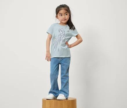 Ein Mädchen steht auf einem Holzsockel. Sie trägt blaue Jeans und ein hellblaues T-Shirt mit einem Aufdruck von Elsa aus Die Eiskönigin.