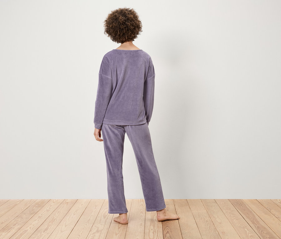 Frau mit lockigem Haar steht mit dem Rücken zum Betrachter und trägt einen lilafarbenen Samtpyjama auf einer hellen Holzoberfläche.