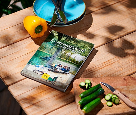 Auf einem Holztisch liegen das Buch »Campingküche«, eine gelbe Paprika, Teller und geschnittene Zucchini.