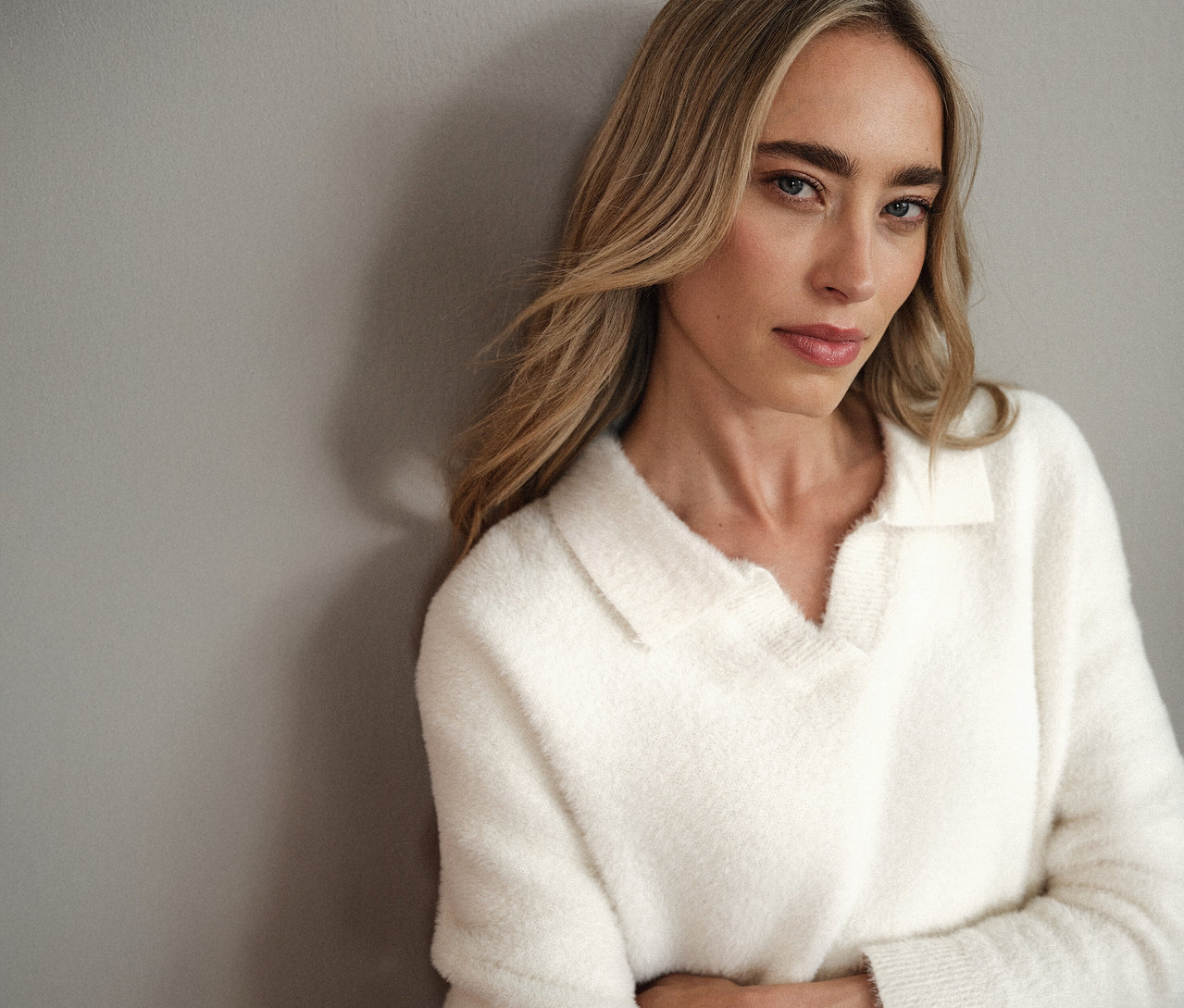 Frau mit blonden Haaren, die einen weißen Street One Strickpullover mit Polokragen trägt und an einer grauen Wand lehnt.