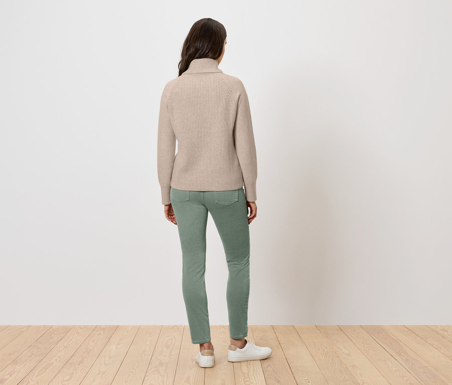 Eine Frau steht mit dem Rücken zur Kamera und trägt einen beigen Rollkragenpullover, grüne Jeans und weiße Turnschuhe.