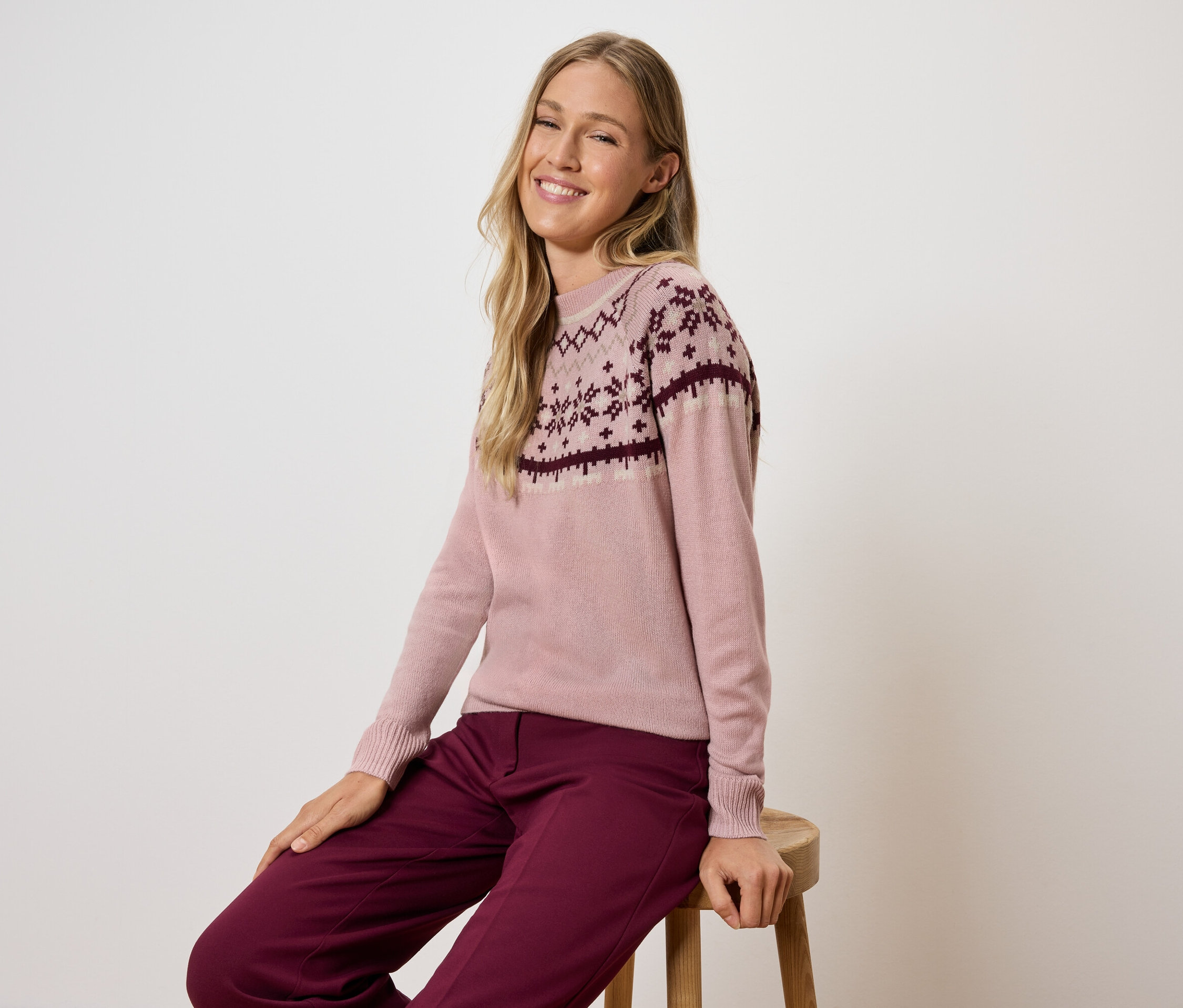 Frau sitzt auf einem Hocker und trägt einen rosa Pullover mit Muster und bordeauxfarbene Hosen.