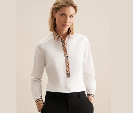 Blonde Frau trägt eine weiße Seidensticker Popeline-Bluse mit Karodetail an Kragen und Manschetten und schwarze Hose, Hände in den Taschen.