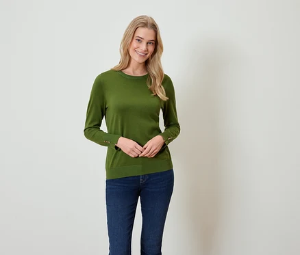 Eine blonde Frau steht und trägt einen grünen Pullover und blaue Jeans.