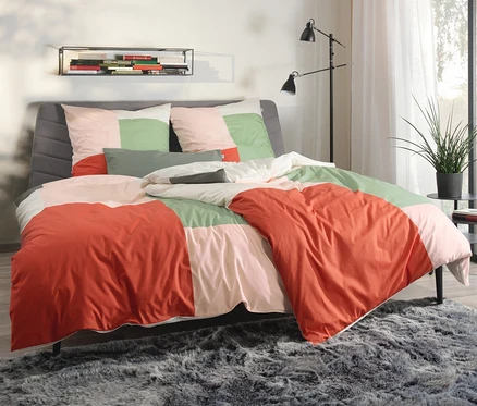 Ein Bett mit rosa-grüner SCHÖNER WOHNEN-Kollektion Bettwäsche und einem grauen Kopfteil. Neben dem Bett steht ein Tisch mit einer Pflanze.