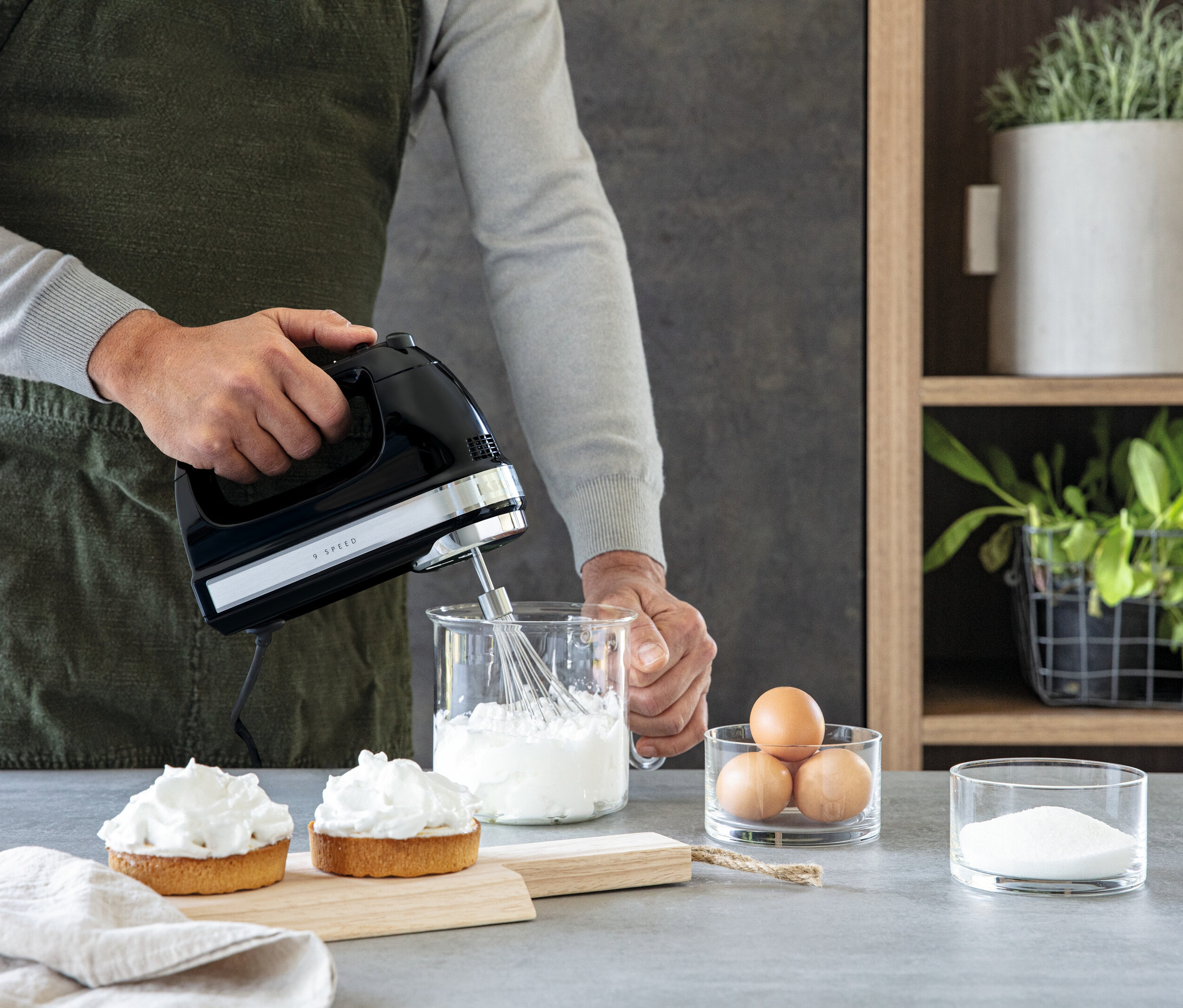 Mann mixt eine weiße Mischung in einer Schüssel mit einem schwarzen Mixer. Auf dem Tisch stehen auch Eier, Zucker und zwei Kuchen.