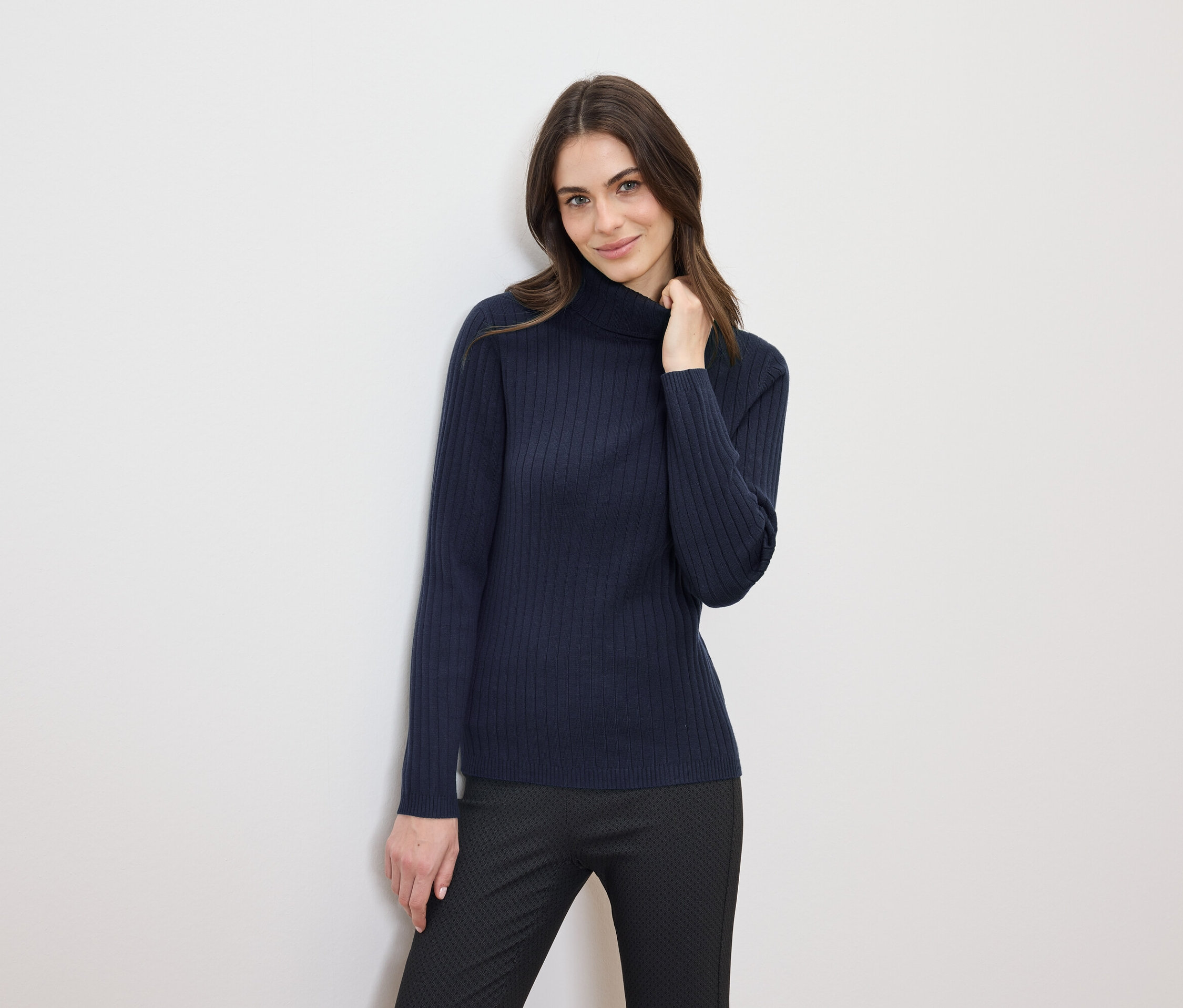 Frau mit braunen Haaren posiert in einem dunkelblauen Rollkragenpullover und einer schwarzen Hose.