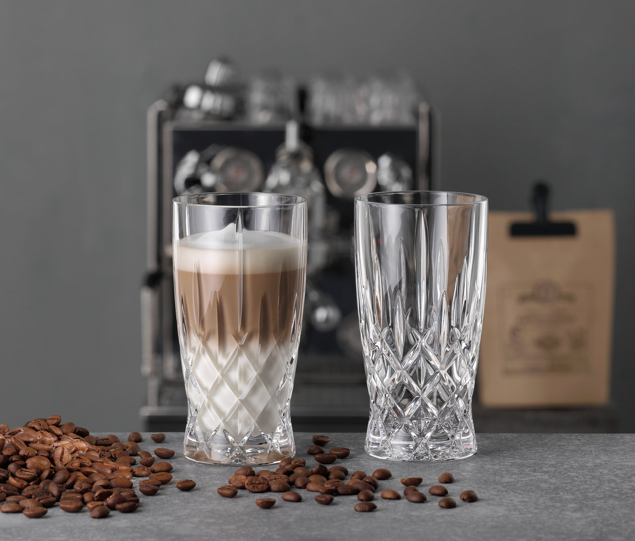 Zwei Nachtmann Latte-Macchiato-Gläser »Noblesse«, eines mit Latte Macchiato, auf einer grauen Oberfläche mit Kaffeebohnen.