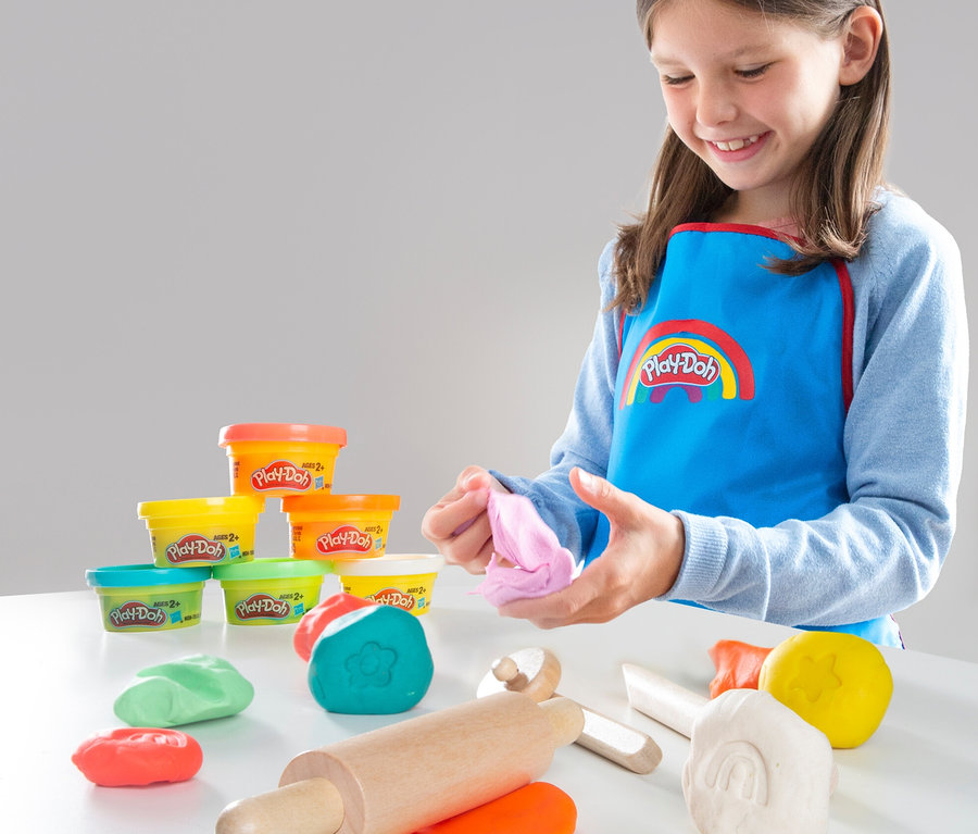 Mädchen mit Play-Doh Schürze spielt mit roba x Play-Doh Knet-Spielset 2 auf einem Tisch.