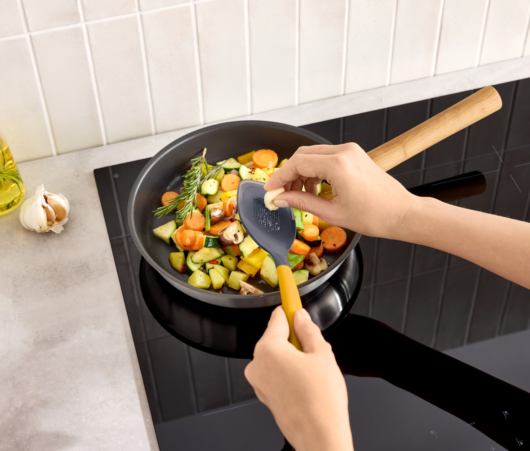 Eine Person reibt eine Knoblauchzehe in eine Pfanne mit Gemüse auf einem Kochfeld.
