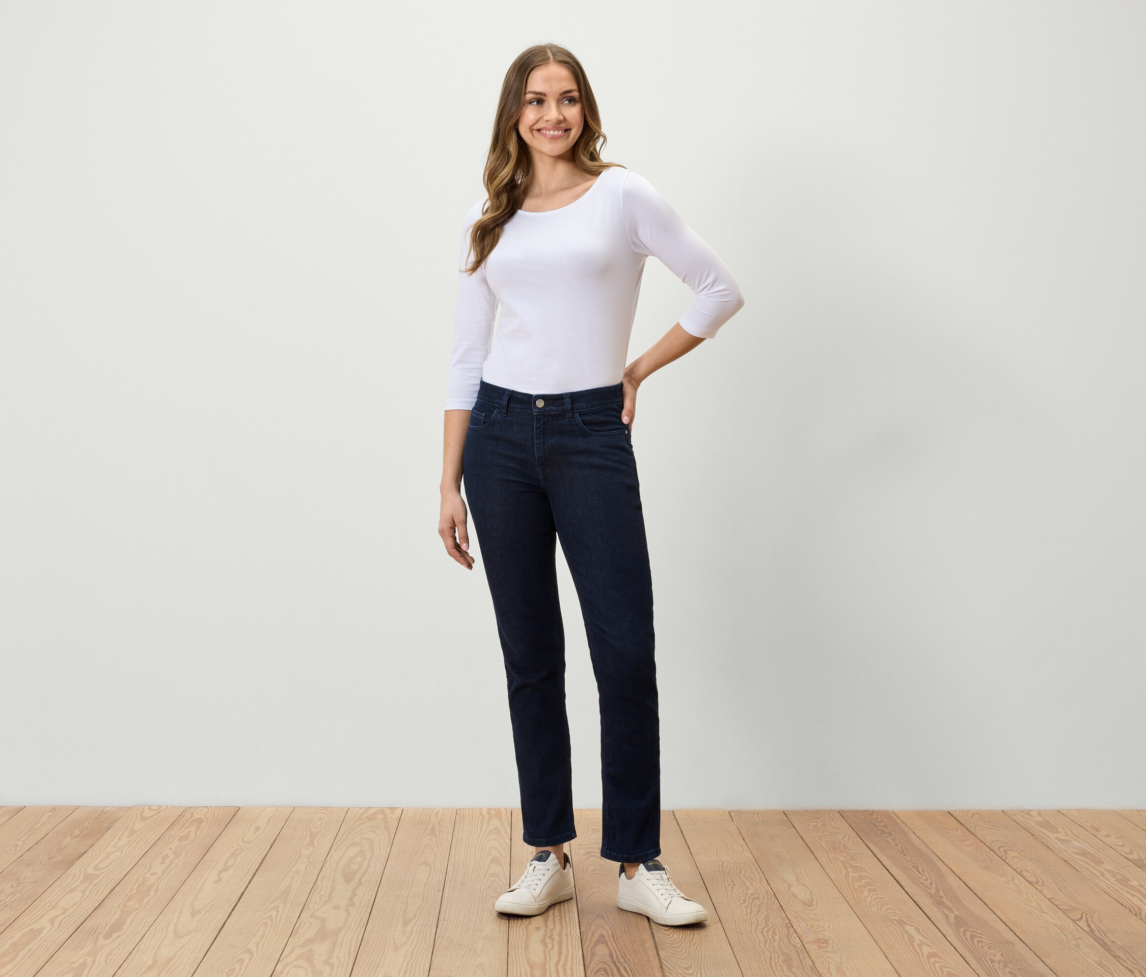 Eine Frau steht auf einem Holzboden und trägt ein weißes Dreiviertelarmshirt, dunkelblaue Jeans und weiße Turnschuhe.