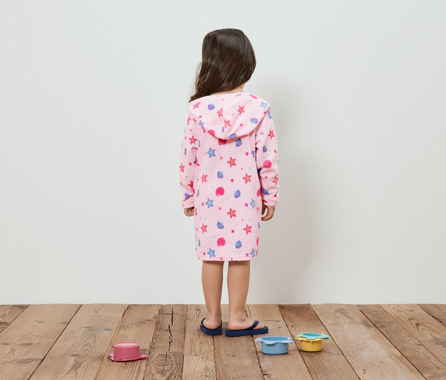 Ein Kind von hinten in einem rosa Bademantel mit Sternen und Muscheln steht auf einem Holzboden neben Spielzeug.