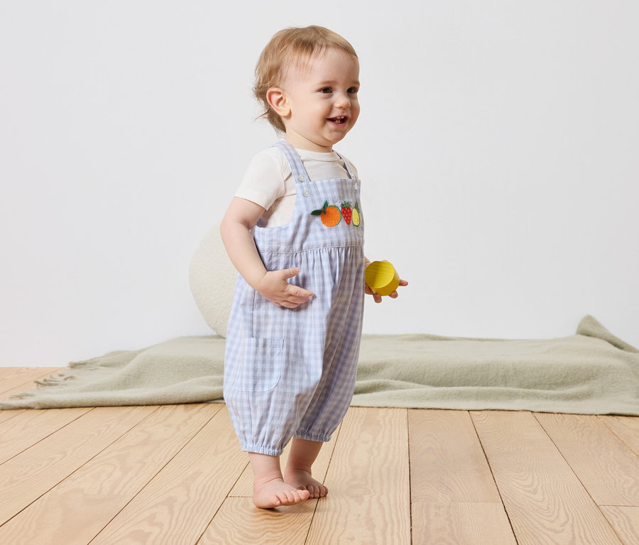 Ein Kleinkind steht barfuß auf einem Holzboden und hält einen gelben Spielzeugblock in der Hand. Das Kind trägt eine blau-weiß karierte Latzhose mit Fruchtmotiven.
