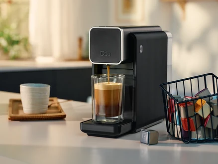 Kaffee läuft aus einer schwarzen Qbo-Kaffeemaschine in ein Glas. Eine Tasse und ein Kapselkorb stehen auf der Küchentheke.