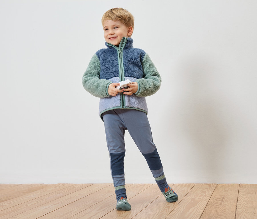 Ein lächelnder Junge mit blonder Kurzhaarfrisur posiert in einer blaugrünen Fleecejacke, blauen Leggings und Socken mit Tiermotiven vor einer Wand.