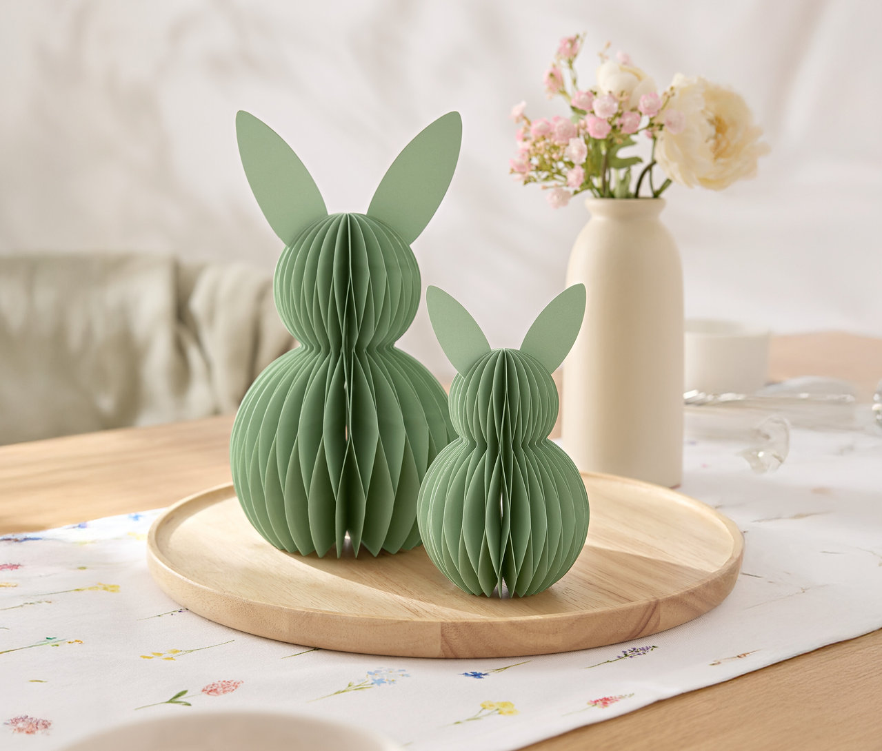 Zwei grüne Papierhasen stehen auf einem Holzteller, dahinter eine Vase mit Blumen.