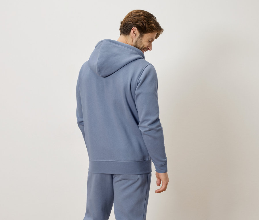 Mann von hinten in blauem Hoodie und Jogginghose vor neutralem Hintergrund.