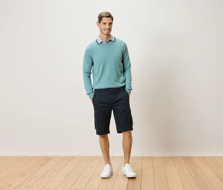 Mann steht mit den Händen in den Taschen vor einer Wand, er trägt einen mintgrünen Pullover, Cargoshorts und weiße Turnschuhe.