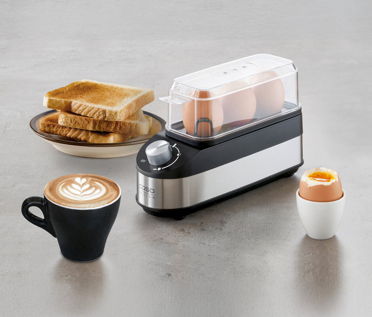 Ein Eierkocher E3 steht neben Toast, Kaffee und einem gekochten Ei.