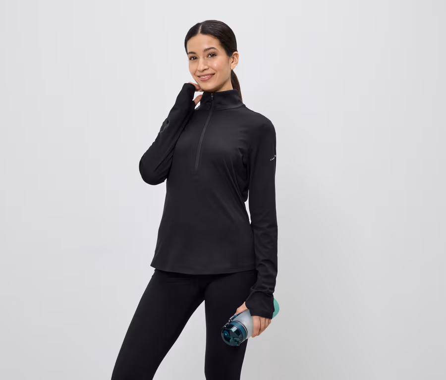 Frau in schwarzem Sportoberteil mit Reißverschluss und Leggings hält eine Wasserflasche vor einem hellen Hintergrund.