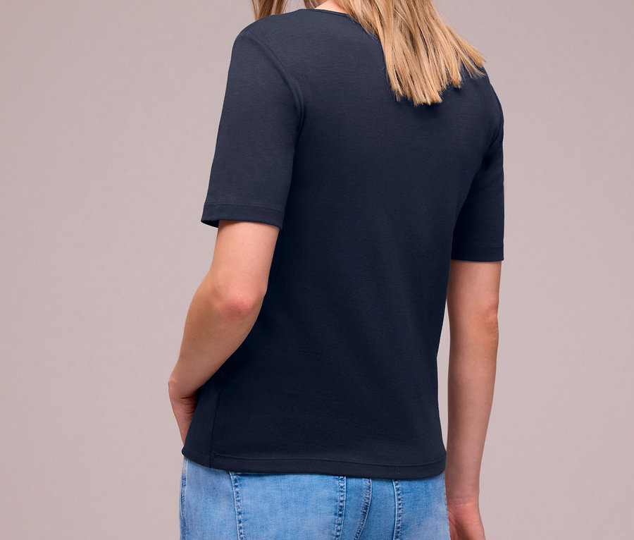 Frau mit langen blonden Haaren, von hinten leicht gedreht, trägt ein navy Street One Kurzarmshirt und hellblaue Jeans, hält eine silberne Jacke.