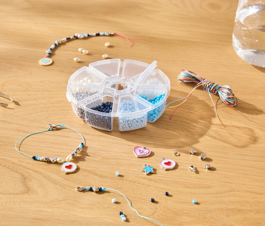 Auf einem Holztisch liegen eine Pinzette, eine Dose mit Perlen, bunte Fäden, eine Wasserflasche und Schmuckstücke.
