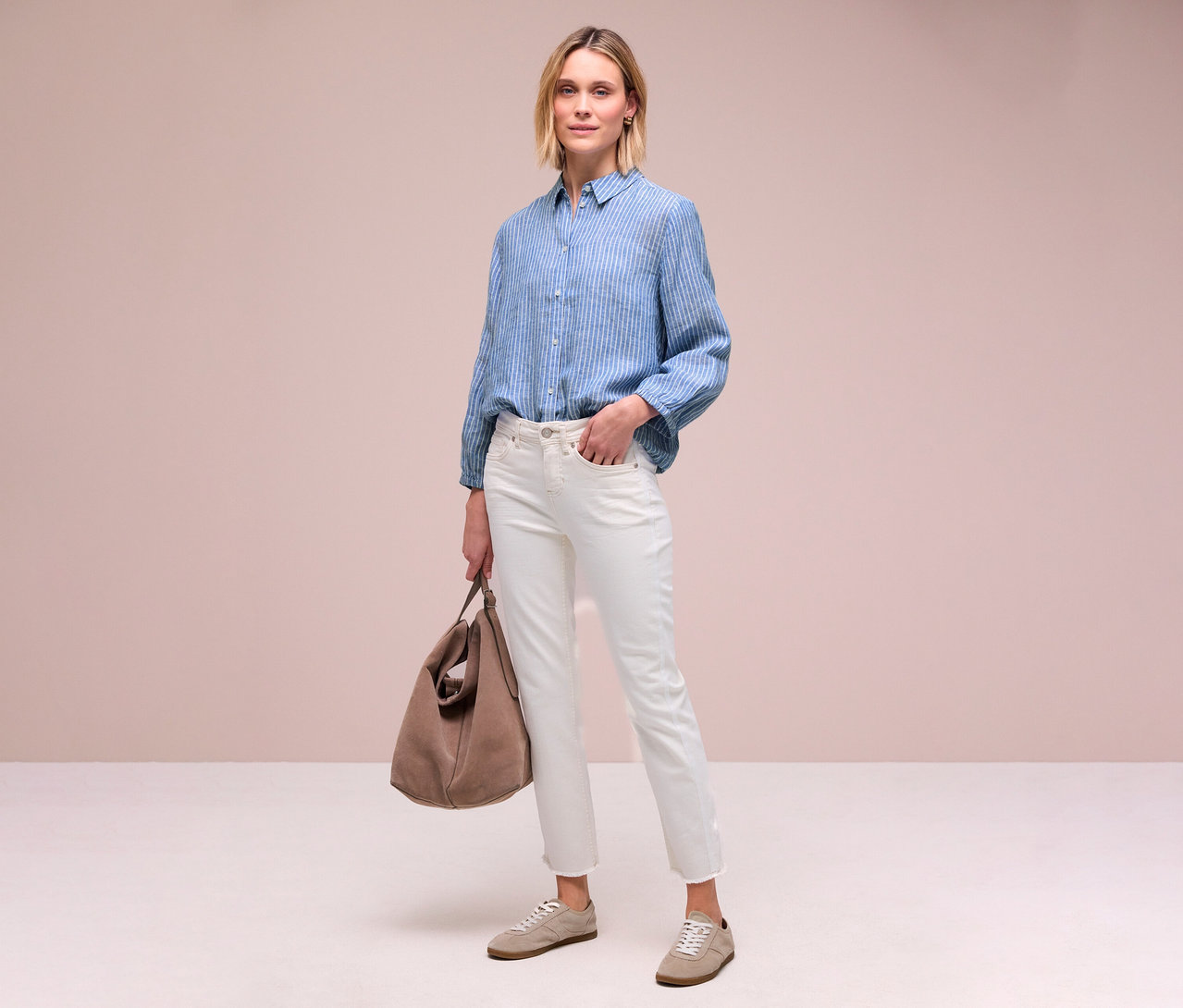 Frau in blau gestreifter Bluse, weißen Jeans, hellbraunen Schuhen und Tasche, stehend.