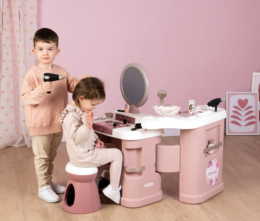 Ein Junge hält einen Spielzeug-Haartrockner über den Kopf eines Mädchens, das auf einem Hocker vor dem Smoby My Beauty Center Kosmetikstudio sitzt.