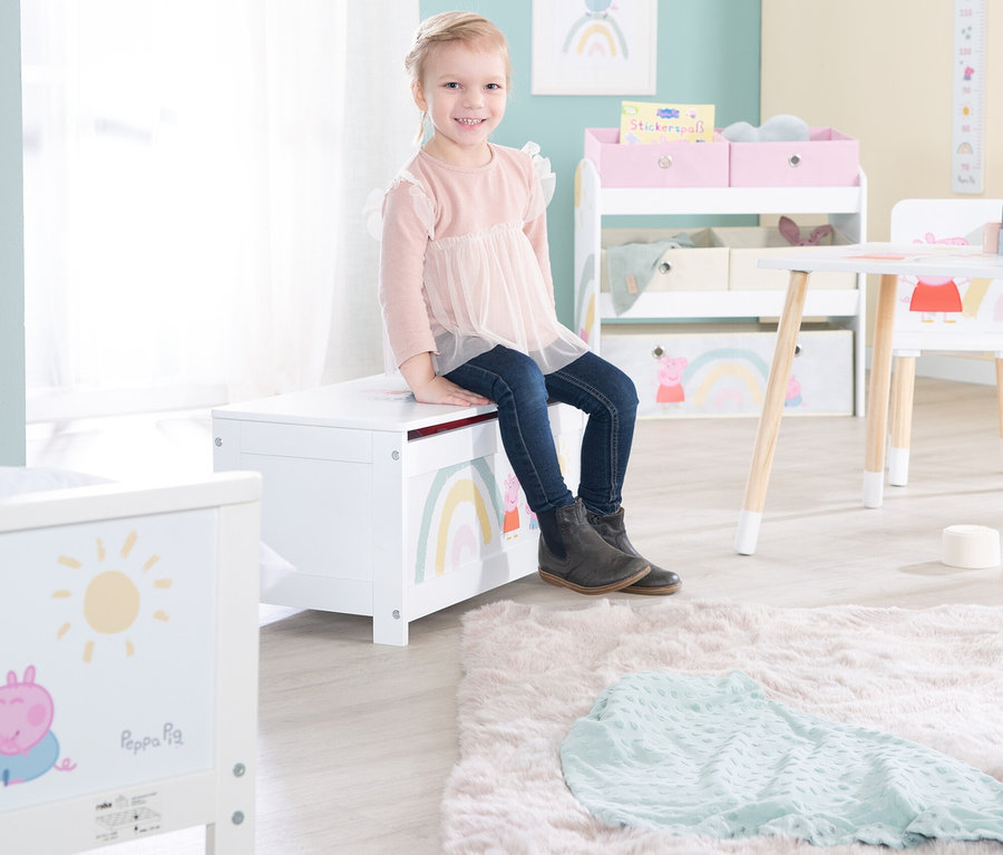 Ein Mädchen sitzt auf einer weißen Spielzeugkiste mit Regenbogen- und Peppa Pig-Motiven.