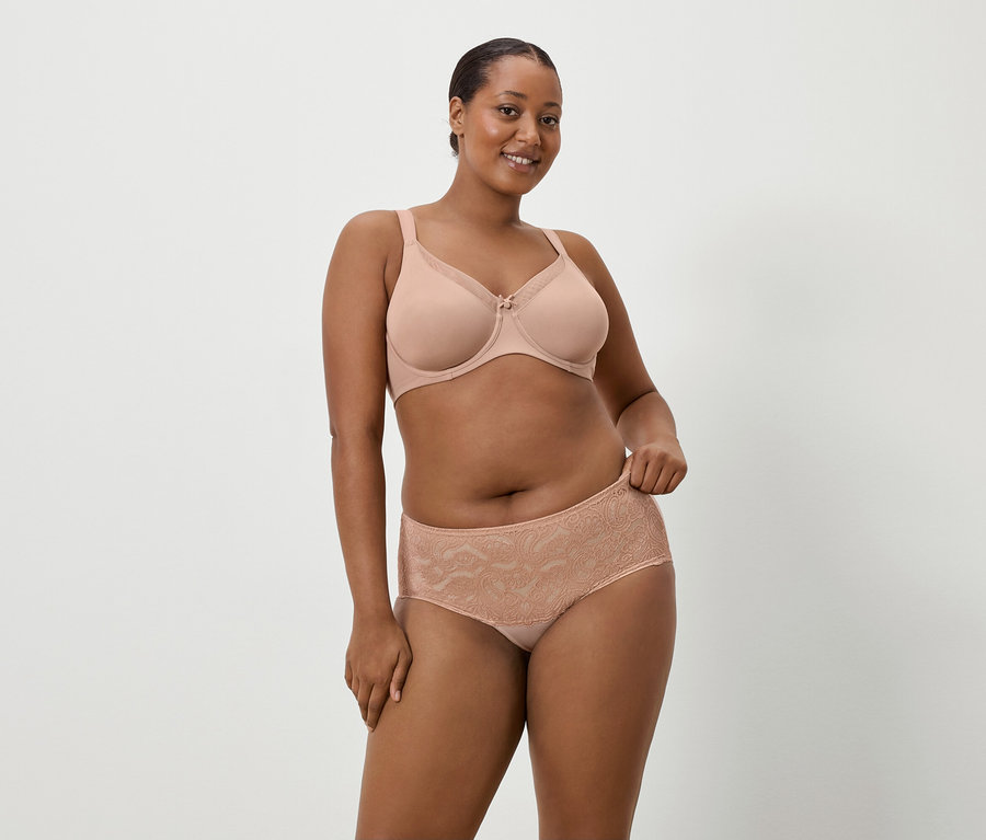 Eine Frau mit dunkler Haut trägt einen nudefarbenen BH und passende Spitzenslips vor einem schlichten Hintergrund.