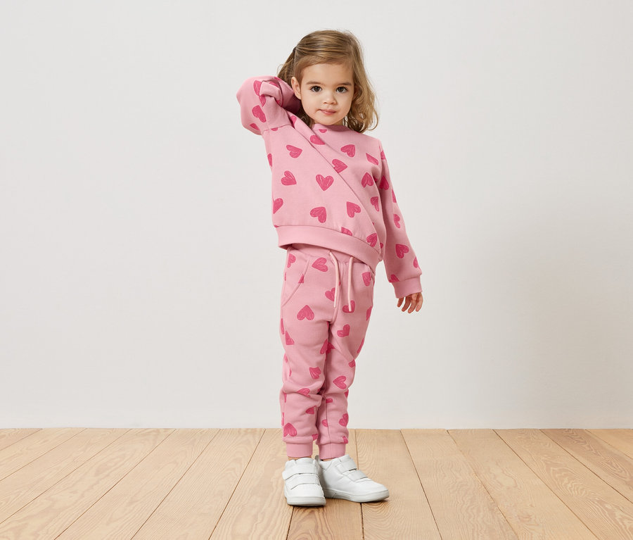 Mädchen mit blonden Haaren trägt einen rosafarbenen Pullover und eine Hose mit Herzmuster und weiße Turnschuhe.